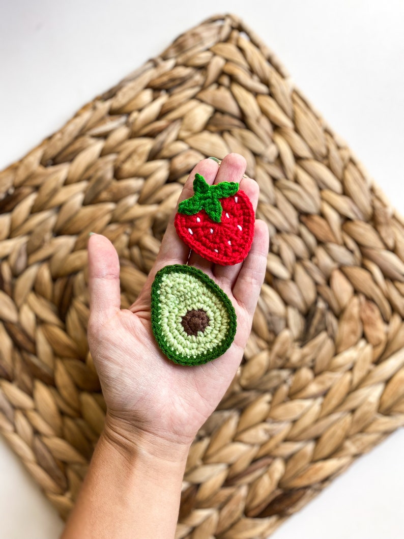May include: A hand holding two crocheted food items: a red strawberry and a green avocado. The items are on a woven straw background.