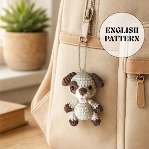 May include: A crocheted dog charm with brown and white details hangs from a silver chain attached to a beige backpack. The dog has brown ears, spots, and paws, with a pink tongue. The image includes a circular graphic that says "ENGLISH PATTERN".