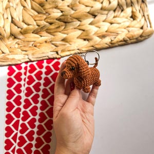 May include: A brown crocheted dachshund keychain with a silver key ring. The keychain is being held by a hand with a red and white bracelet.