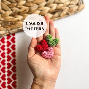 Puede incluir: Tres corazones tejidos a crochet en diferentes colores: rojo, rosa y verde. Los corazones se sostienen en una mano, con el texto "ENGLISH PATTERN" visible en el fondo.