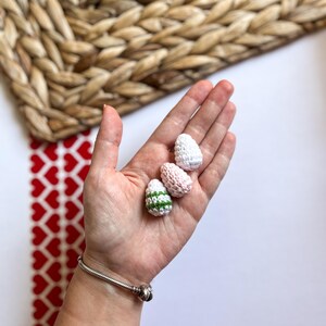 Puede incluir: Tres huevos de Pascua de ganchillo en una mano. Un huevo es blanco, otro es rosa y blanco, y el &uacute;ltimo es blanco con rayas verdes.
