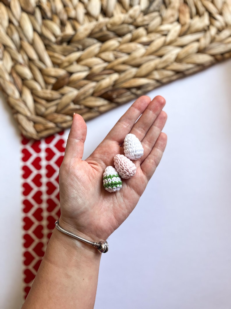 Puede incluir: Tres huevos de Pascua de ganchillo en una mano. Un huevo es blanco, otro es rosa y blanco, y el &uacute;ltimo es blanco con rayas verdes.