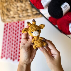 Puede incluir: Un peque&ntilde;o juguete de jirafa de crochet con manchas marrones y una cara blanca. La jirafa est&aacute; siendo sostenida por las manos de una persona.
