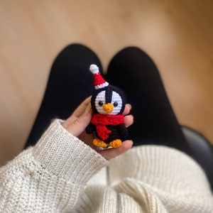 May include: A small crocheted penguin wearing a red Santa hat and a red scarf. The penguin is being held in someone's hand.