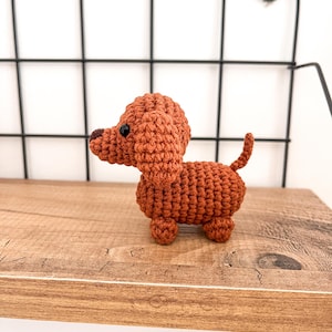 May include: A small, crocheted dachshund toy in a brown color. The toy is sitting on a wooden shelf.