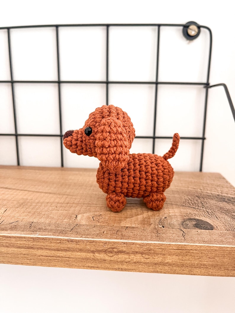 May include: A small, crocheted dachshund toy in a brown color. The toy is sitting on a wooden shelf.