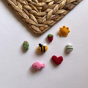 Puede incluir: Una colecci&oacute;n de peque&ntilde;os art&iacute;culos tejidos a crochet, que incluyen una abeille, un coraz&oacute;n, un pez, un cactus, una fresa, un sol y un huevo. Los art&iacute;culos est&aacute;n hechos con hilo de diferentes colores y est&aacute;n dispuestos sobre un fondo blanco.