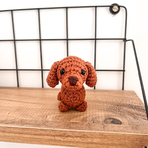 May include: A small, crocheted brown dog toy with black eyes and a brown nose. The toy is sitting on a wooden shelf.
