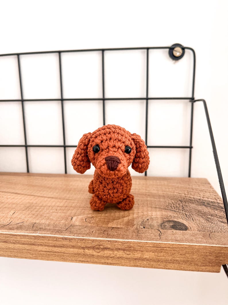 May include: A small, crocheted brown dog toy with black eyes and a brown nose. The toy is sitting on a wooden shelf.