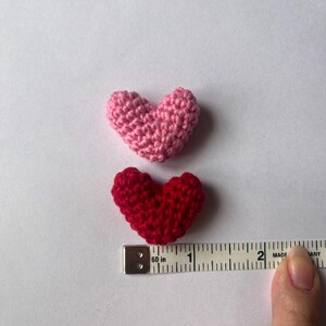 May include: Two crocheted hearts, one pink and one red, are shown next to a measuring tape. The hearts are approximately 1 inch in size.