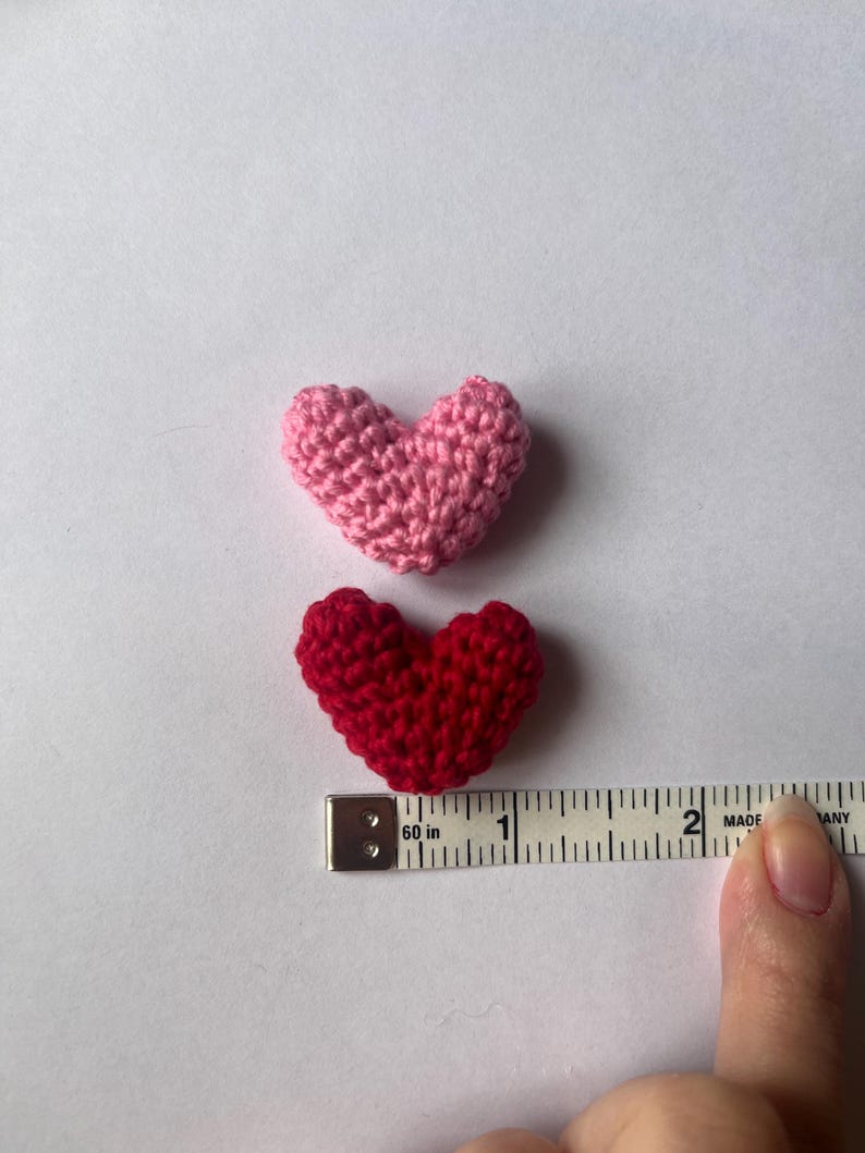 May include: Two crocheted hearts, one pink and one red, are shown next to a measuring tape. The hearts are approximately 1 inch in size.
