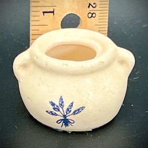May include: A small, off-white ceramic pot with two handles and a blue floral design. The pot is approximately 1 inch in diameter and has a rustic, aged appearance. The pot is likely a decorative item or a miniature container.