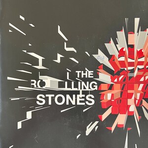 Könnte beinhalten: Ein schwarzes Poster mit dem Text THE ROLLING STONES in weißen Großbuchstaben. Rechts davon scheint ein stilisiertes rot-weißes Grafikelement nach außen zu explodieren, wobei scharfe, geometrische Splitter über den dunklen Hintergrund strahlen.