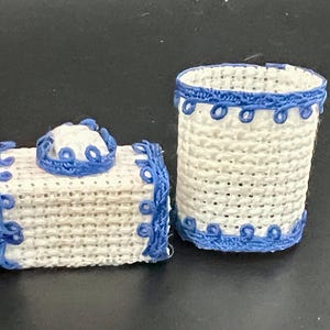 May include: Two miniature woven containers. One is rectangular with a lid, the other is cylindrical. Both are white with blue trim and decorative accents. The objects are likely decorative or for small items.