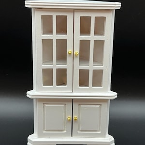 May include: A white miniature cabinet with glass-paned doors and gold-colored knobs. The cabinet has a two-door bottom section and a top section with four glass panes per door. The cabinet is designed with a classic, traditional style.
