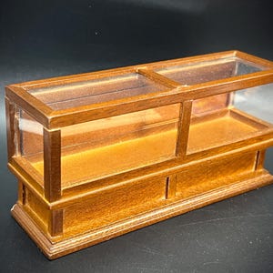 May include: A miniature wooden display case with a glass top and sides. The case has a warm, brown finish and features two separate display areas. The bottom section has two small, vertical indentations. The overall design is reminiscent of a vintage shop display.