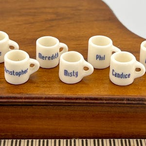 Pode incluir: Sete canecas de cerâmica branca com letras azuis. As canecas são etiquetadas com os nomes Joyce, Christopher, Meredith, Phil, Misty, Candice e Marily.