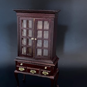 May include: A dark brown wooden cabinet with glass doors and three drawers. The cabinet has a traditional design with cabriole legs and brass hardware.