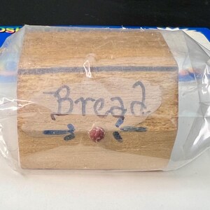 Peut inclure: Une petite boîte en bois avec le mot "Bread" écrit sur une étiquette transparente. La boîte est enveloppée dans du plastique transparent. Un petit élément décoratif rouge est en dessous du mot "Bread".