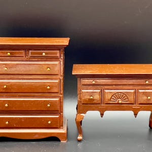 Puede incluir: Dos muebles de madera en miniatura, una cómoda y una mesa auxiliar, ambos en un rico acabado marrón. La cómoda tiene múltiples cajones con pomos de latón. La mesa auxiliar presenta un diseño de sol.