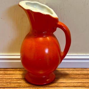 Könnte beinhalten: Ein kleiner, orangefarbener Keramik-Krug mit weißem Rand. Der Krug hat einen runden Körper und einen kleinen Henkel.