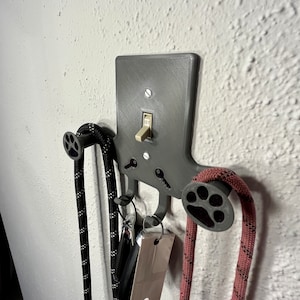 Könnte beinhalten: Eine 3D-gedruckte Wandhalterung mit einem Lichtschalter und zwei Haken in Form von Pfoten. Zwei Leinen hängen an den Haken.