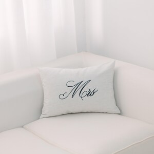 Op de afbeelding: Een wit kussen met het woord "Mrs." geborduurd in zwarte cursieve letters.