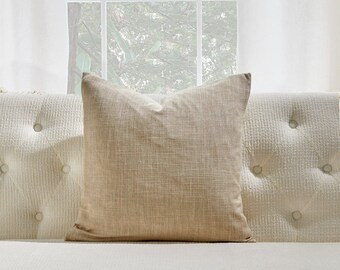 May include: A beige throw pillow with a subtle textured fabric. The pillow is placed on a white tufted sofa.