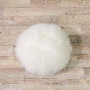 May include: A white fluffy round sheepskin rug with long, soft fur. The rug is on a light brown wood floor.