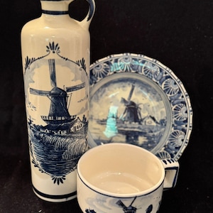 May include: A collection of Delftware pottery with blue and white windmill designs. The set includes a tall, handled bottle, a plate, and a teacup. The pottery has a traditional, hand-painted appearance, with intricate details of windmills and landscapes.