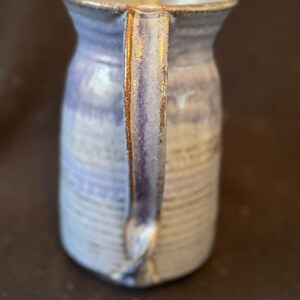 May include: A handmade ceramic jug with a light blue glaze and a brown handle. The jug has a wide mouth and a curved handle. The exterior features horizontal lines. This rustic jug is perfect for serving beverages.
