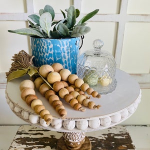 Può includere: Un porta torte a due livelli bianco con una base in legno. Il livello superiore è decorato con perline bianche e presenta un mazzo di perline di legno, un vaso macchiato di blu e bianco con una pianta verde e una campana di vetro con uova di Pasqua verdi e bianche.