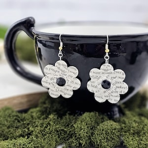 May include: A pair of book page flower earrings with black beads. The earrings are hanging from a black teacup.