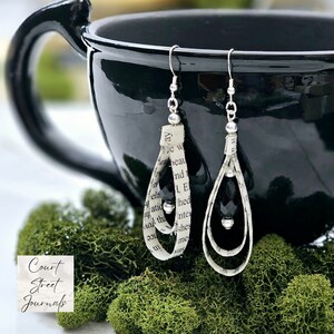 May include: A pair of dangle earrings made with book pages and silver beads. The earrings are hanging from a black mug.