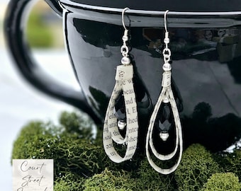 May include: A pair of dangle earrings made with book pages and silver beads. The earrings are hanging from a black mug.