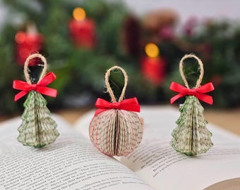 May include: Three handmade Christmas ornaments. Two are tree-shaped, and one is round. They are made from book pages and have red ribbon bows and twine hangers. The ornaments are displayed on an open book.