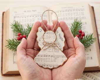 May include: A decorative angel ornament crafted from vintage sheet music paper with gold glitter edges, featuring a circular head, wing-shaped body, and a small twine bow at the neck, suspended by a thin twine loop for hanging purposes.