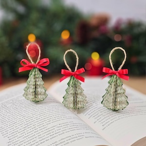 May include: Three handmade Christmas tree ornaments made from book pages, with green edges and red ribbon bows. Each tree has a natural twine loop for hanging. The ornaments are displayed on an open book.