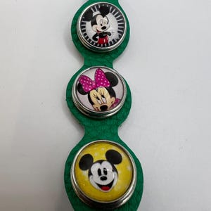 Puede incluir: Pulsera de cuero verde con tres colgantes circulares con personajes de Disney. El colgante superior muestra a Mickey Mouse, el colgante central a Minnie Mouse y el colgante inferior a Mickey Mouse. Cada colgante tiene un borde plateado.