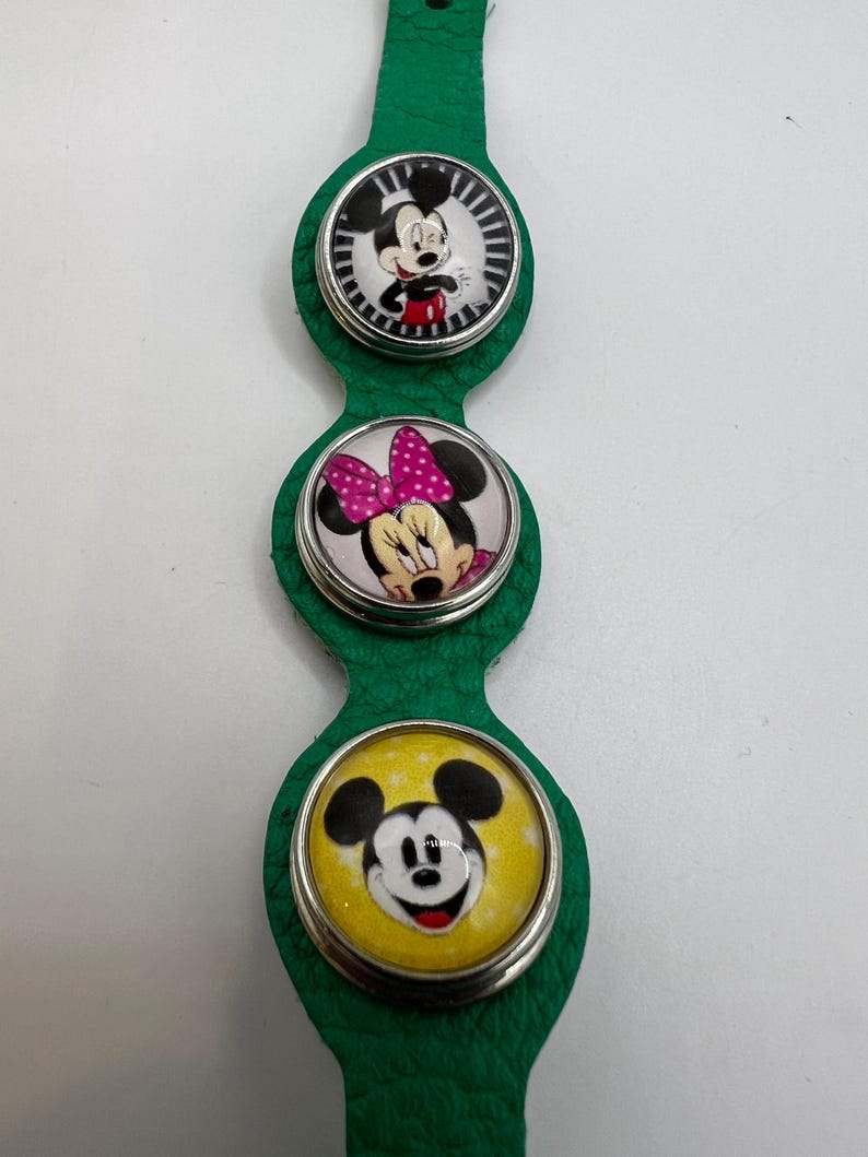 Puede incluir: Pulsera de cuero verde con tres colgantes circulares con personajes de Disney. El colgante superior muestra a Mickey Mouse, el colgante central a Minnie Mouse y el colgante inferior a Mickey Mouse. Cada colgante tiene un borde plateado.