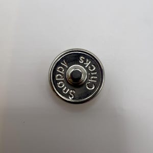 Puede incluir: Un cierre magn&eacute;tico plateado con las palabras "Snappy Chicks" grabadas en el borde. El cierre tiene un bot&oacute;n central en relieve sobre un fondo blanco.