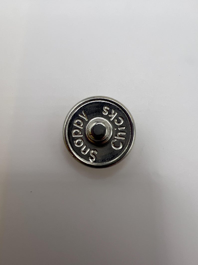 Puede incluir: Un cierre magn&eacute;tico plateado con las palabras "Snappy Chicks" grabadas en el borde. El cierre tiene un bot&oacute;n central en relieve sobre un fondo blanco.