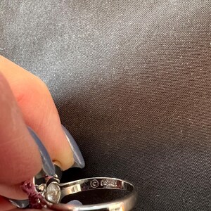 May include: A silver ring featuring a clear gemstone and small red stones. The ring is stamped with the Disney logo. The ring is held by a person with blue nail polish.