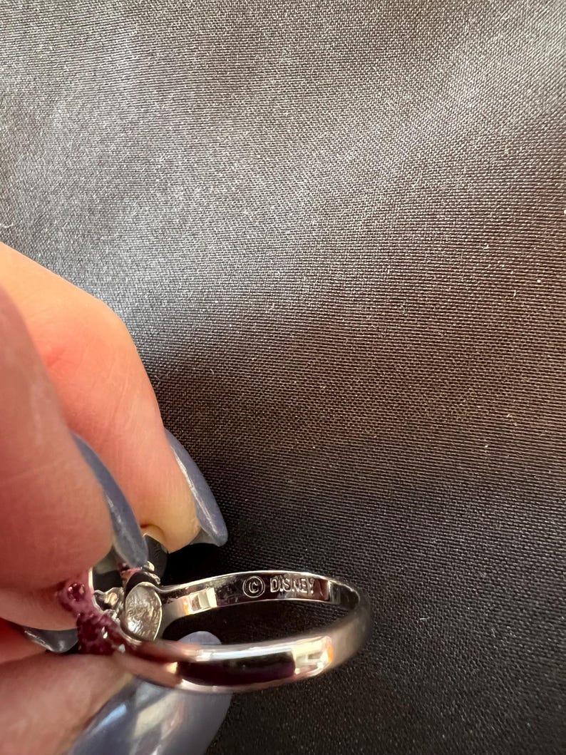 May include: A silver ring featuring a clear gemstone and small red stones. The ring is stamped with the Disney logo. The ring is held by a person with blue nail polish.
