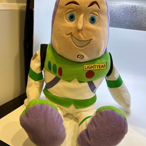 May include: A plush toy of Buzz Lightyear from the Toy Story franchise. The toy is sitting down with its arms at its sides. The toy is wearing a white and green spacesuit with a purple chest piece. The toy has a purple head with a yellow face and blue eyes. The toy has a name tag that says "LIGHTYEAR".