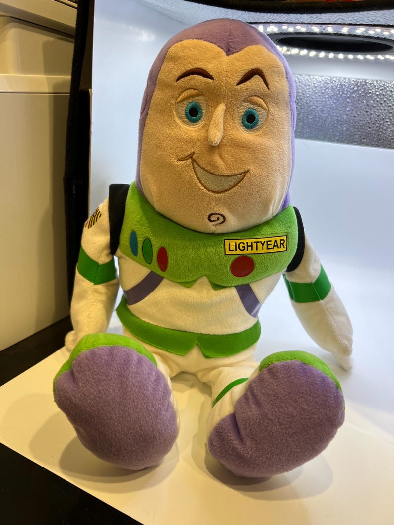 May include: A plush toy of Buzz Lightyear from the Toy Story franchise. The toy is sitting down with its arms at its sides. The toy is wearing a white and green spacesuit with a purple chest piece. The toy has a purple head with a yellow face and blue eyes. The toy has a name tag that says "LIGHTYEAR".