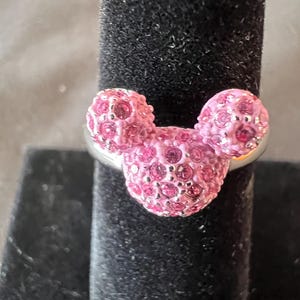 May include: A silver ring with a Mickey Mouse design. The ring features three pink, crystal-covered spheres. The crystals sparkle. The ring is displayed on a black velvet surface, creating a contrast with the bright pink crystals.