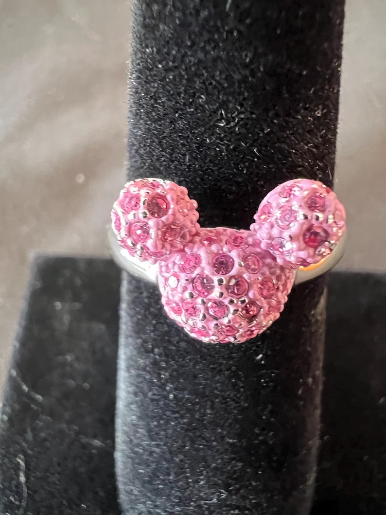 May include: A silver ring with a Mickey Mouse design. The ring features three pink, crystal-covered spheres. The crystals sparkle. The ring is displayed on a black velvet surface, creating a contrast with the bright pink crystals.