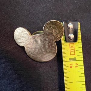 Pu&ograve; includere: Una spilla dorata a forma di Topolino con una superficie strutturata. La spilla &egrave; accanto a un metro a nastro giallo, che mostra una misura di circa 2,5 cm.