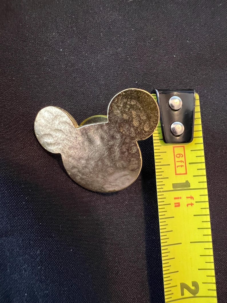 Pu&ograve; includere: Una spilla dorata a forma di Topolino con una superficie strutturata. La spilla &egrave; accanto a un metro a nastro giallo, che mostra una misura di circa 2,5 cm.